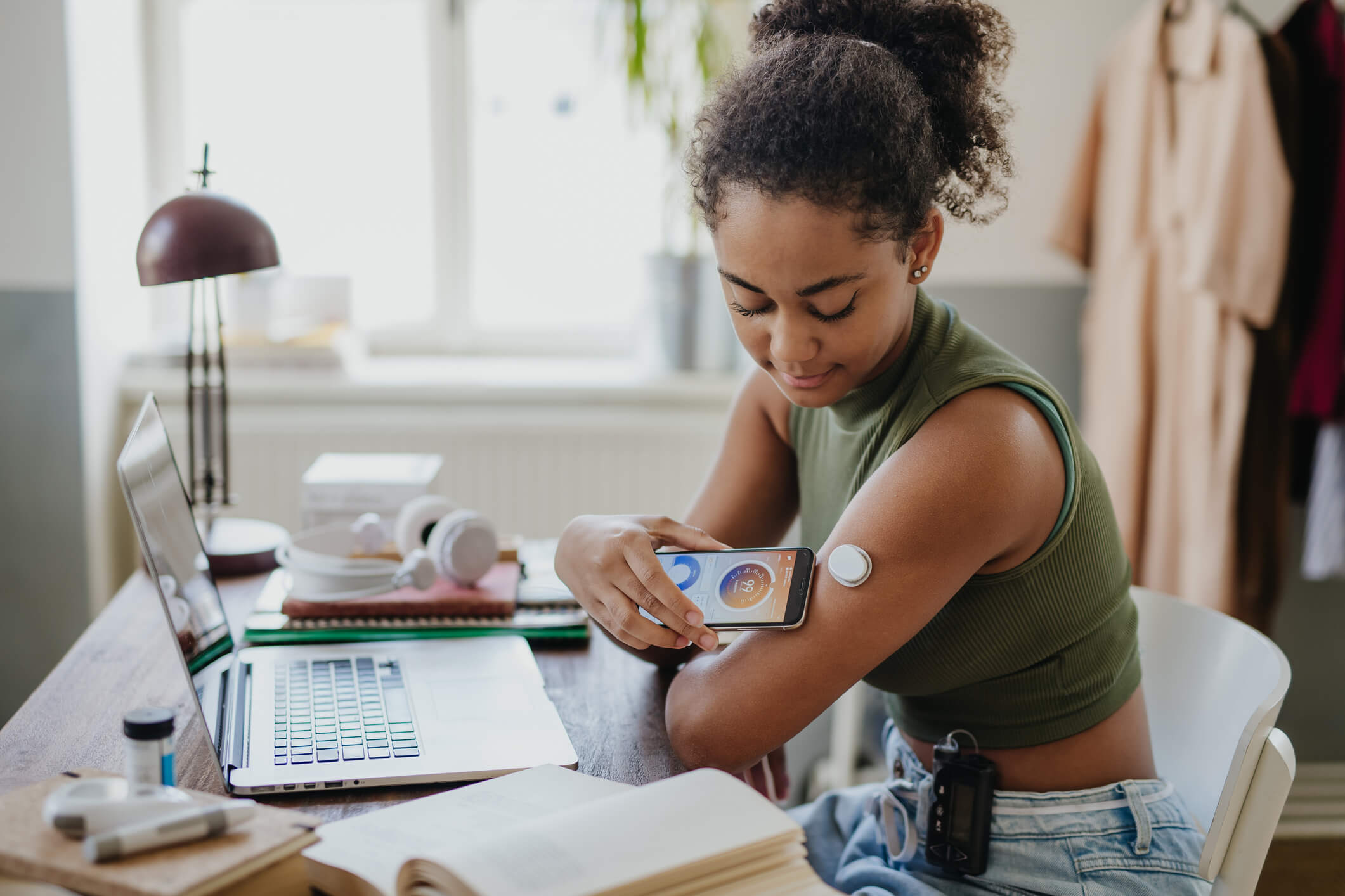 Teenage girl connecting smartphone to continuous glucose monitor, checking blood glucose during day. Student doing routine blood check while studying. Girl’s CGM sensor monitoring blood sugar levels in real time. Concept of daily life with chronic illness, diabetes for teenagers.